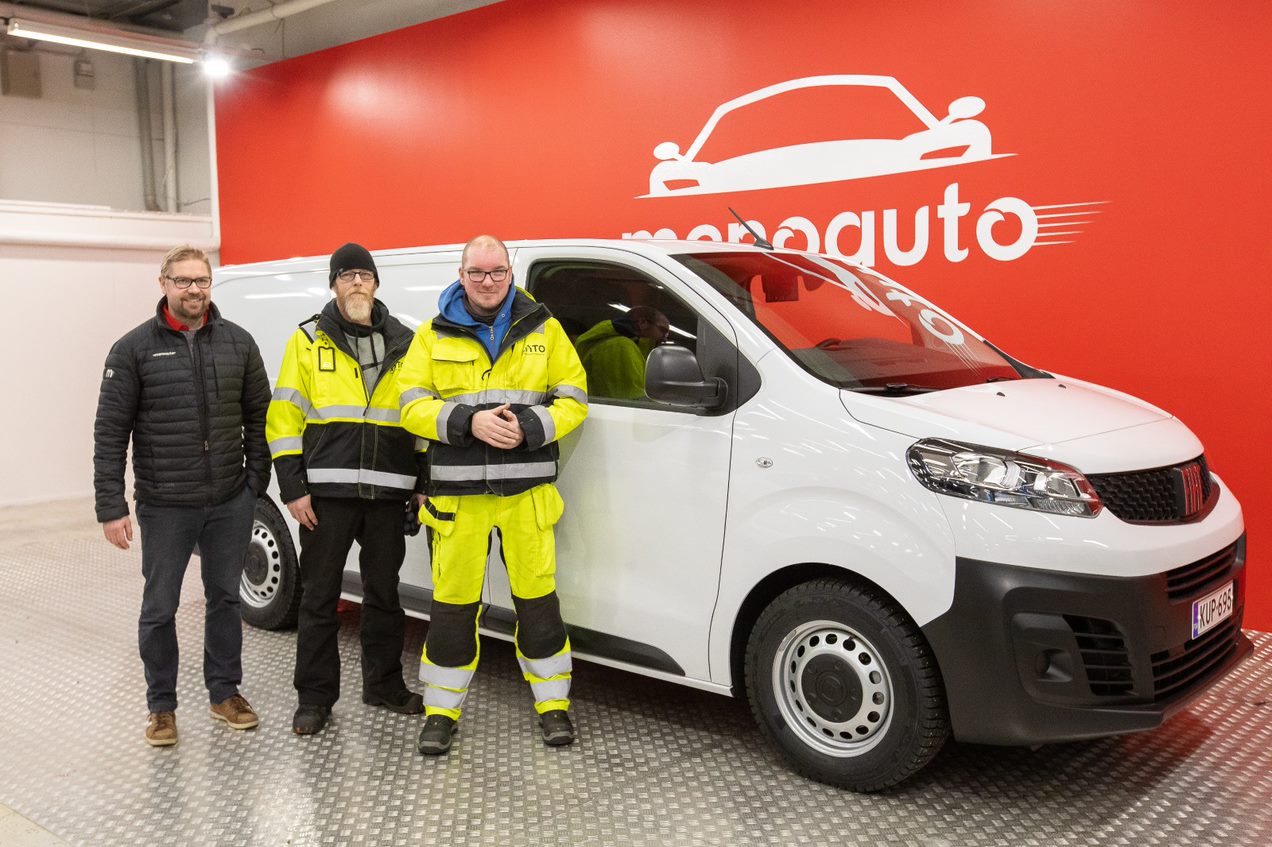 Kolme miestä seisoo valkoisen pakettiauton edessä, taustalla oranssi seinä, jossa lukee ”menoauto” ja kuva auton keulasta.